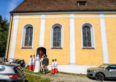 Kirchliche Trauung in St. Alban, Diessen am Ammersee, Brautpaar vor gelber Kirche in Hochzeitskleidung.