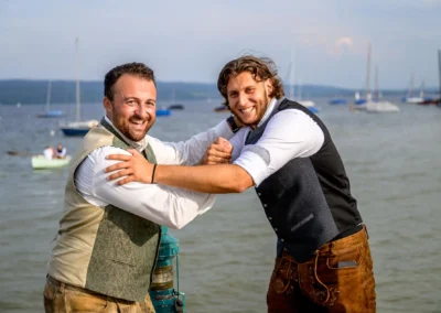 Zwei fröhliche Männer in traditioneller Tracht auf einem Steg beim Seehaus Riederau am Ammersee zur Hochzeit.