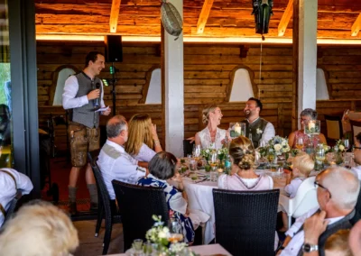 Hochzeitsfeier mit Rede, Brautpaar lacht am Tisch, Hochzeitsreportage in traditioneller Umgebung.