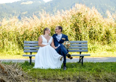 Brautpaar beim Fotoshooting in Bad Wiessee am Tegernsee, romantisch auf einer Bank sitzend vor Bergkulisse.