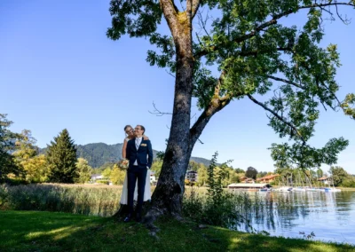 Brautpaar am Tegernsee, Bad Wiessee, unter Baum mit See und Bergen als Kulisse. Hochzeitsreportage-Fotografie.