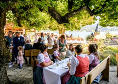 Hochzeitsfeier im Terrassenhof Bad Wiessee mit Gästen in traditioneller Tracht unter Bäumen am Seeufer.