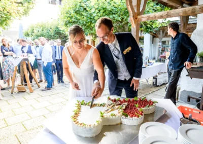 Brautpaar schneidet Hochzeitstorte bei Feier im Terrassenhof Bad Wiessee an. Gesellige Stimmung im Freien.
