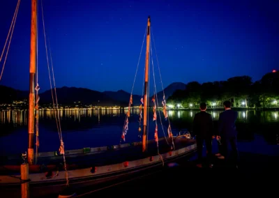 Hochzeitsfeier im Bootshaus Terrassenhof Bad Wiessee am Abend, romantische Seeansicht mit Gästen und Segelboot.