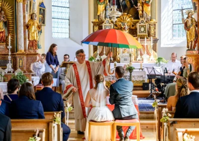 Kirchliche Trauung in St. Margaretha, Pfaffing. Priester segnet Brautpaar unter Regenbogenschirm vor Altar.
