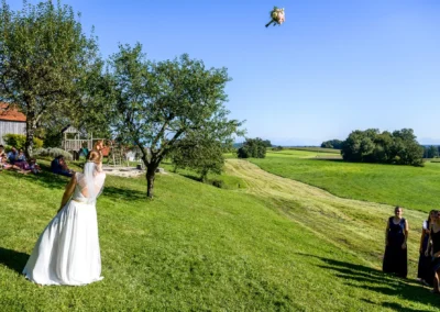 Braut wirft Blumenstrauß bei Hochzeit auf Landgut Kugler-Alm, Ebersberg, mit Gästen im Hintergrund.