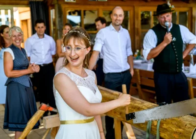 Brautpaar bei traditionellem Baumstammsägen auf Hochzeit im Landgut Kugler-Alm, Ebersberg, nahe München.