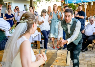 Brautpaar zersägt Baumstamm bei Hochzeit auf der Kugler-Alm in Ebersberg nahe München. Gäste applaudieren.
